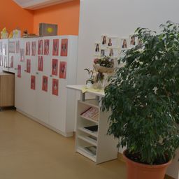 Ein Innenraum mit orangefarbener Wand hat einen Schrank mit Fotos, einen Tisch mit Pflanze und Büchern und ein Regal mit Blumenarrangement und einer Box.