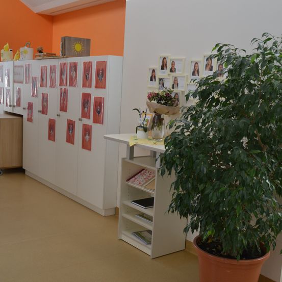 Ein Innenraum mit orangefarbener Wand hat einen Schrank mit Fotos, einen Tisch mit Pflanze und Büchern und ein Regal mit Blumenarrangement und einer Box.