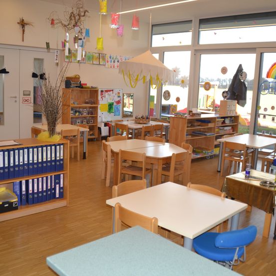 Ein leerer Klassenraum mit Holztischen, Stühlen und Regalen. Dekorationen und Kunstwerke an den Wänden, große Fenster.