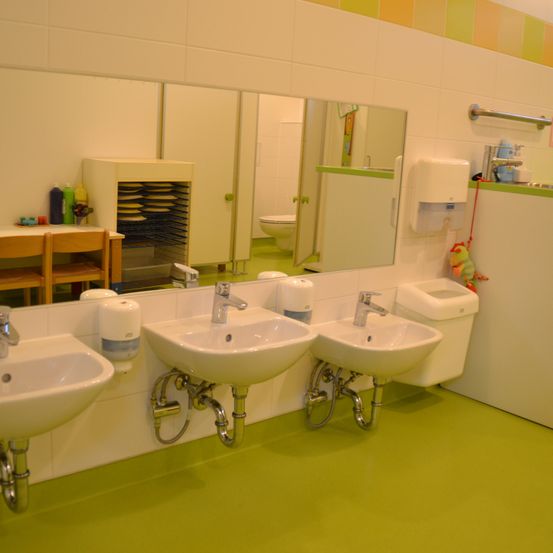 Ein Badezimmer mit drei Waschbecken, einer Toilette und einem Handtrockner. Es gibt auch Regale mit Reinigungsprodukten.