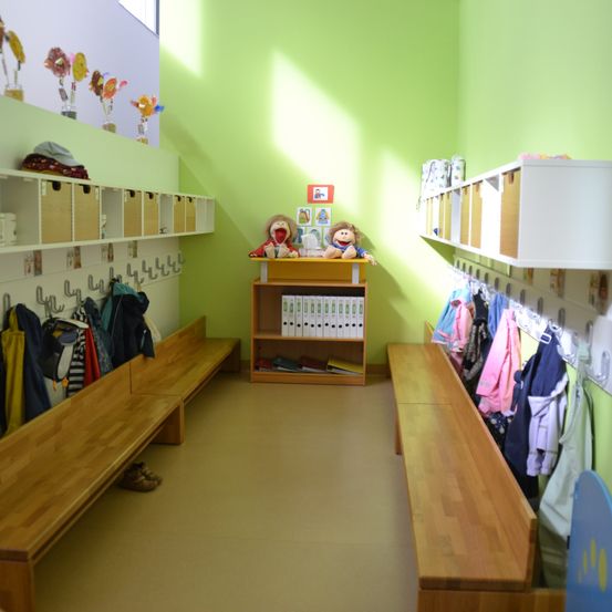 Ein Raum mit grünen Wänden hat zwei Holzbänke, ein kleines Regal und hängende Kleidung. Der Raum hat Bücher, Spielzeug und Aufbewahrungsboxen auf den Regalen.