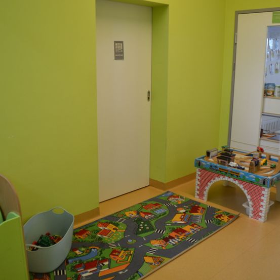 Ein Kinderspielzimmer mit einem Eisenbahn-Set auf einem Spielteppich, einem Spielzeug-Aufbewahrungsbehälter und einer gelben Wand.