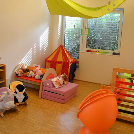 Ein Kinderspielzimmer mit Holzboden, Spielzeug und Möbeln. Ein Bett mit Kuscheltieren, ein rosa Stuhl und ein Zelt. Ein orangefarbener Stuhl und ein gelber Hängesessel. Ein Fenster mit Jalousien.