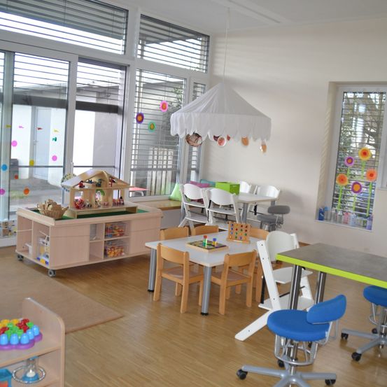 Ein heller, sauberer Raum mit Holzboden und großen Fenstern, ausgestattet mit einem kleinen Hausspielset, Tischen mit Stühlen und bunten Dekorationen an den Wänden.