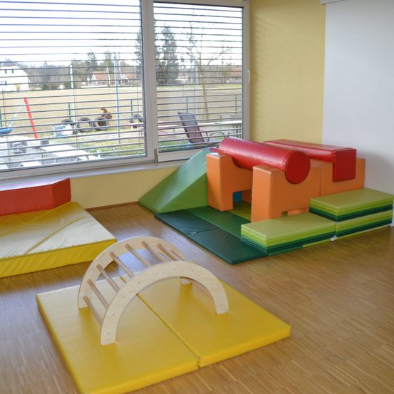 Ein Spielzimmer mit Holzboden und verschiedenen farbigen Matten und Kissen. Ein hölzerner Bogen ist auf einer gelben Matte platziert. In der Nähe des Fensters gibt es einen Blick auf einen Spielplatz im Freien.