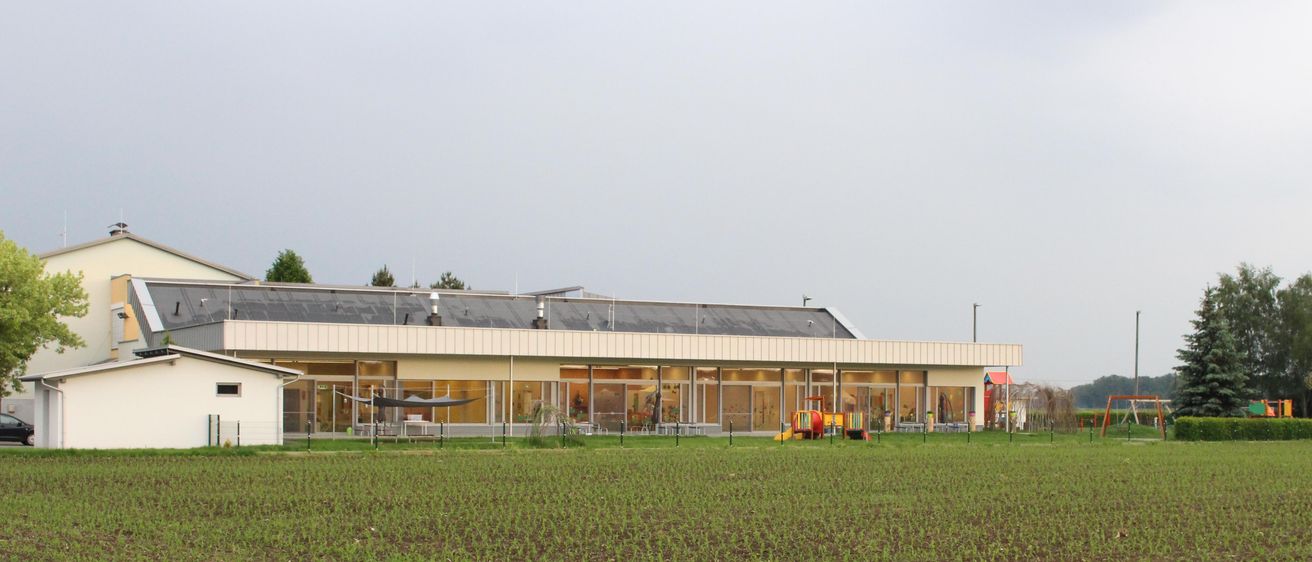 Ein großes Gebäude mit einem Spielplatz davor, umgeben von einem Feld unter einem bewölkten Himmel.