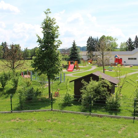 Ein Kinderspielplatz mit einer roten Rutsche und verschiedenen Spielgeräten auf einem grünen Rasenfeld mit einem Gebäude im Hintergrund.