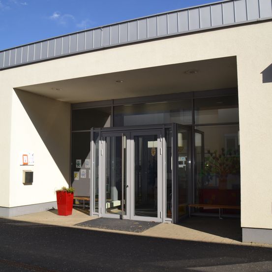 Der Eingang eines modernen Gebäudes mit einer Glastür und einem Fenster, einem roten Pflanzgefäß mit Pflanzen und einer Bank rechts.