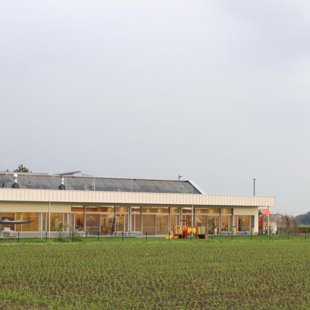Ein Gebäude mit großen Fenstern und einem Spielplatz steht mitten auf einem breiten grünen Feld unter einem bewölkten Himmel.