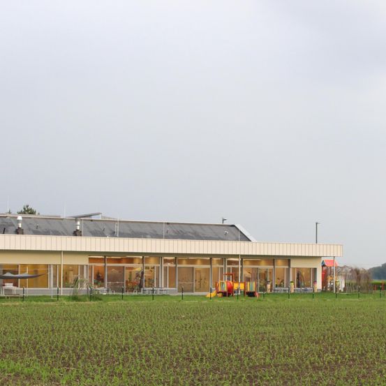 Ein Gebäude mit großen Fenstern und einem Spielplatz steht mitten auf einem breiten grünen Feld unter einem bewölkten Himmel.