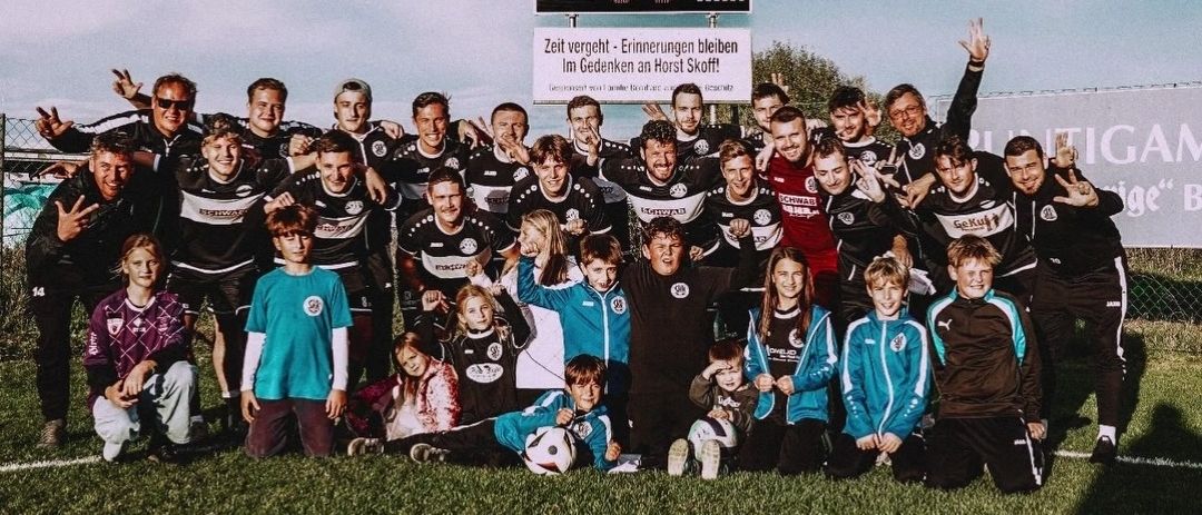Eine Fußballmannschaft mit Spielern und Unterstützern posiert für ein Foto auf einem Rasenfeld, wobei ein Anzeigetafel eine Zeit von 18:88 im Hintergrund anzeigt.