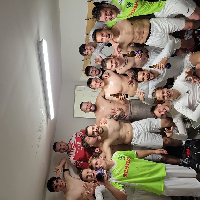 A group of young men in sports gear are posing for a photo in a locker room. Some have tattoos and hold bottles. A light is on the wall.