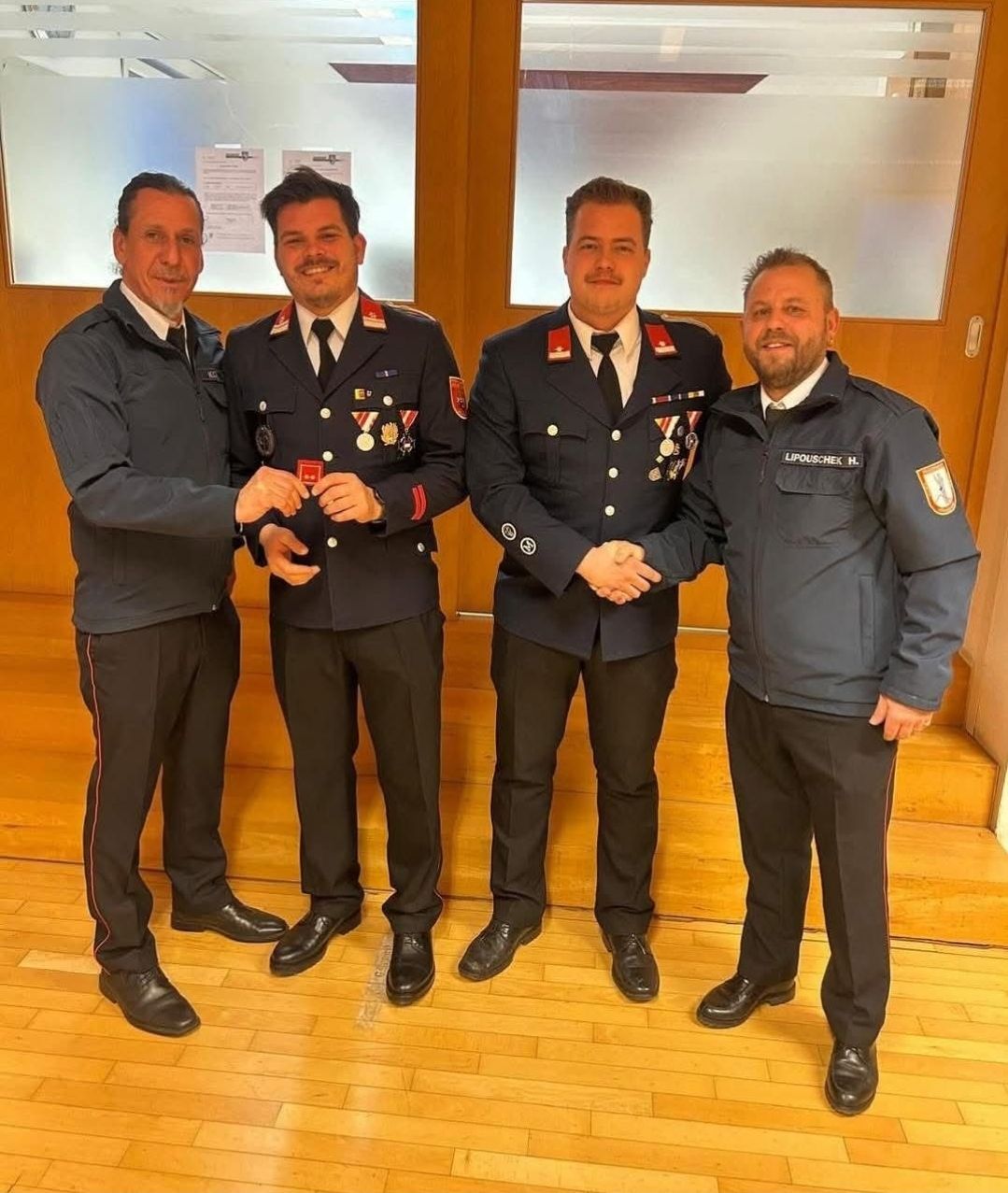 Four men in uniform stand on a wooden floor, two are shaking hands while one is receiving an award.