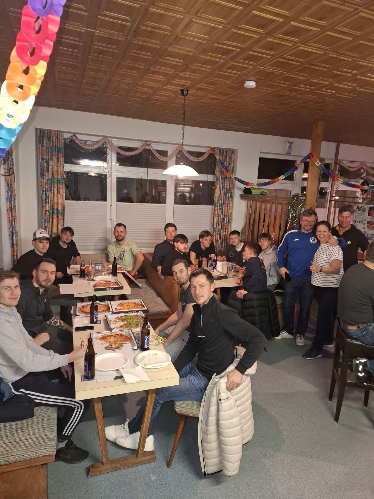 A group of people are gathered in a restaurant, sitting around tables with plates of pizza and beer bottles. Some people are standing behind the tables.