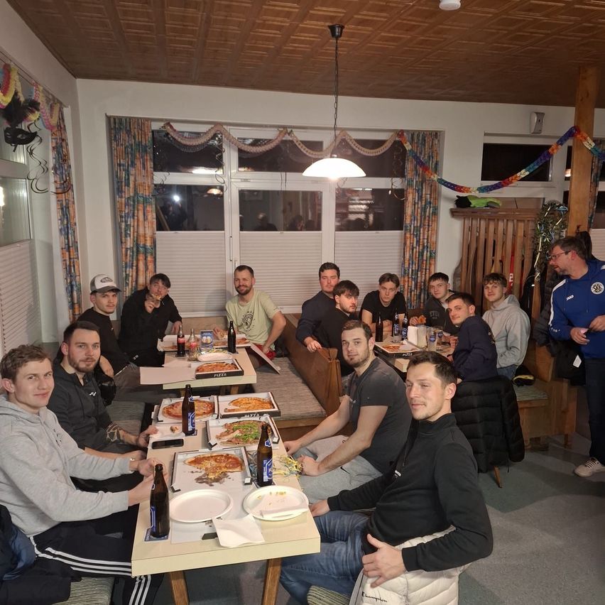 A group of people sit around tables in a restaurant, some with pizza plates and bottles. A man in a blue shirt stands at the back.