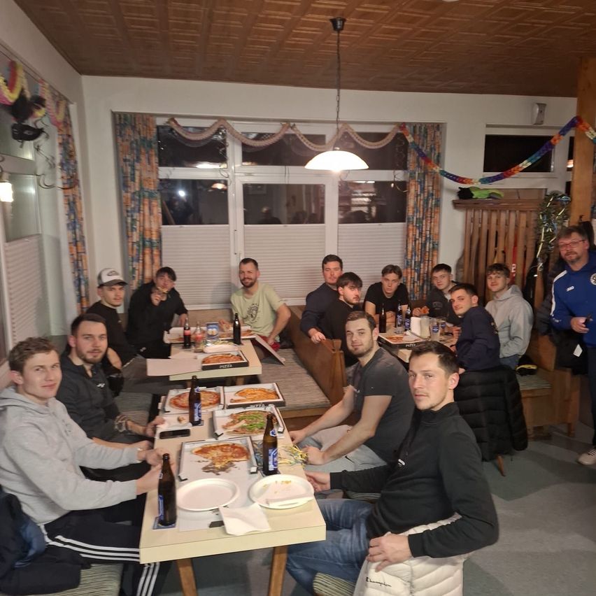 A group of men gathered around a table in a restaurant. Some of them are wearing hats and glasses. Pizza and beer are on the table. The room is decorated with curtains and a hanging lamp.