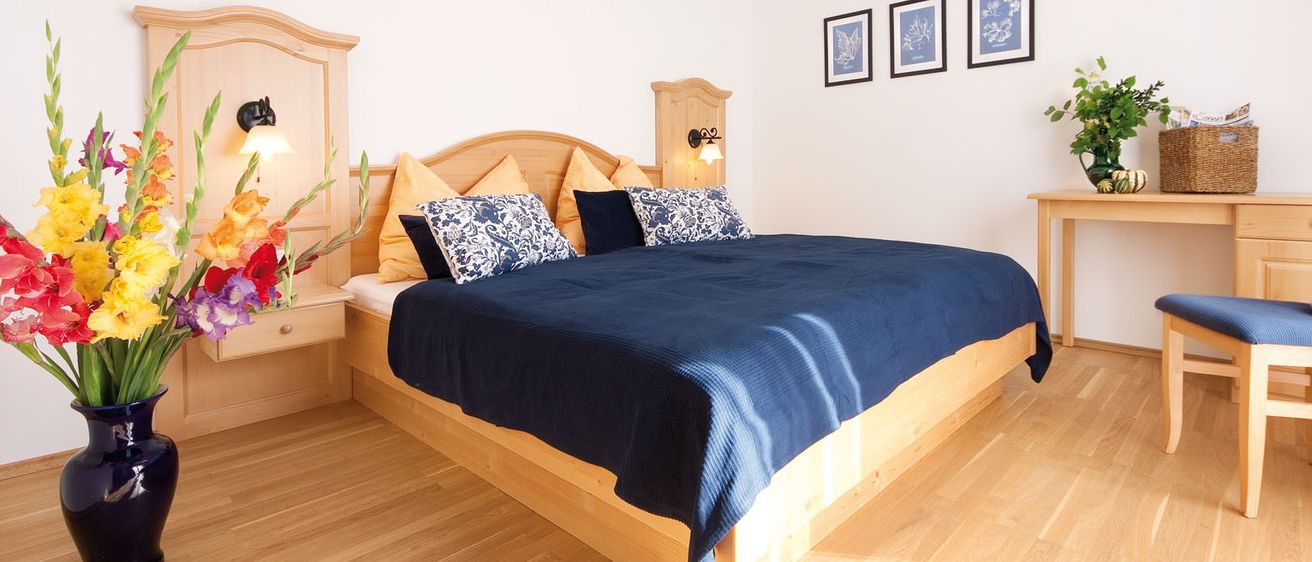 Ein ordentlich eingerichtetes Schlafzimmer mit einem Holzbett, blauer Bettwäsche, dekorativen Kissen und drei gerahmten Drucken an einer weißen Wand. Das Bett hat ein hölzerne Kopfteil, und eine Lampe ist an der Wand über ihm montiert.