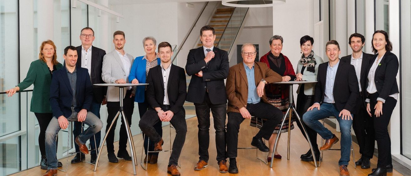 Eine Gruppe von Menschen, geschäftlich gekleidet, versammelt sich in einem modernen Büro. Sie sitzen auf Hockern und stehen um Tische herum und lächeln für ein Foto. Im Hintergrund sind eine Treppe und große Fenster zu sehen.