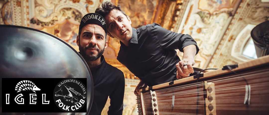Zwei Männer stehen eng beieinander, einer trägt eine Brooklyn-Kappe und der andere spielt Sitar. Sie stehen vor einer verzierten Wand.