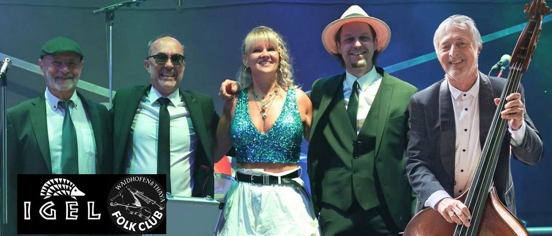 Drei Personen posieren für ein Foto, wobei die Frau in der Mitte ein blaues Pailletten-Top und einen weißen Rock trägt. Der Mann links trägt eine Sonnenbrille und einen Anzug. Der Mann rechts trägt einen grünen Anzug und einen Hut.