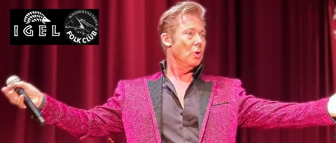 Ein Mann in einem pinken Pailletten-Jacke und schwarzem Hemd steht auf der Bühne und performt mit einem roten Vorhang im Hintergrund.