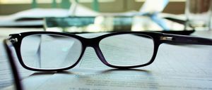 Ein Paar schwarze Brille mit klaren Gläsern liegt auf einem Dokument, möglicherweise einem Vertrag. Das Dokument ist leicht verschwommen und hat etwas darauf geschriebenen Text.
