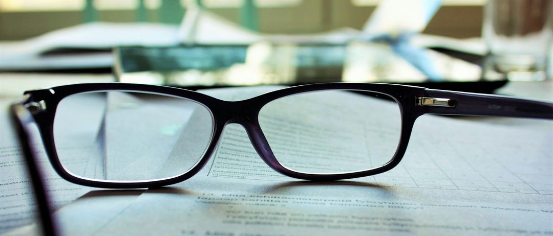 Ein Paar schwarze Brille mit klaren Gläsern liegt auf einem Dokument, möglicherweise einem Vertrag. Das Dokument ist leicht verschwommen und hat etwas darauf geschriebenen Text.