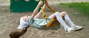 Ein junges Mädchen schwingt auf einem gelben Schaukelpferd in einem Spielplatz. Sie trägt eine weiße Haube, ein graues Kleid und weiße Socken. Im Hintergrund befindet sich eine rote Bank und ein Kinderwagen.