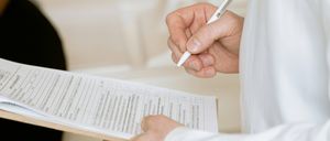 Eine Person in einem weißen Hemd füllt ein Formular mit einem Stift aus. Das Formular befindet sich auf einem Klemmbrett. Der Hintergrund ist unscharf.