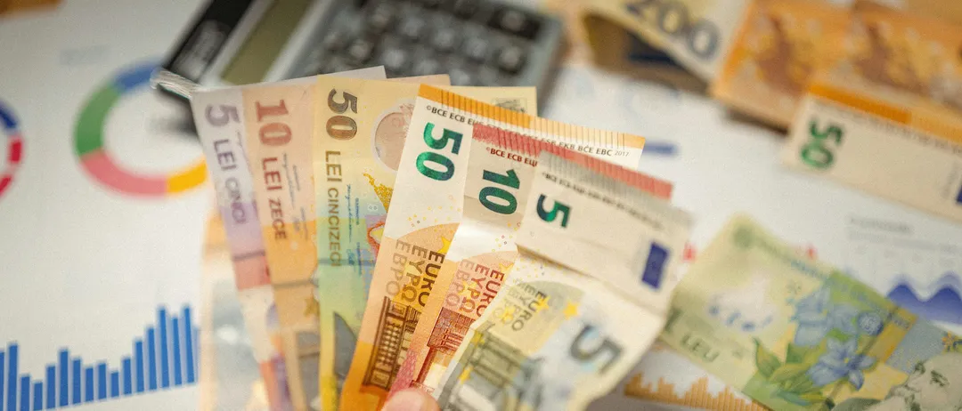 Eine Hand hält eine Sammlung mehrfarbiger Euro-Banknoten, mit einem Taschenrechner und Finanzdiagrammen im Hintergrund.