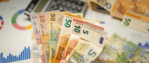 Eine Hand hält eine Sammlung mehrfarbiger Euro-Banknoten, mit einem Taschenrechner und Finanzdiagrammen im Hintergrund.