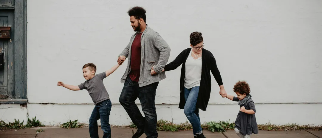 Eine vierköpfige Familie, darunter zwei kleine Kinder, geht fröhlich Hand in Hand auf einem Bürgersteig und lächelt, während sie für ein Foto posiert.