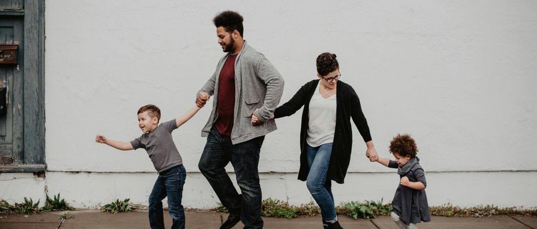 Eine vierköpfige Familie, darunter zwei kleine Kinder, geht fröhlich Hand in Hand auf einem Bürgersteig und lächelt, während sie für ein Foto posiert.
