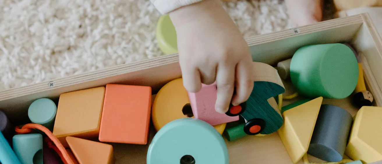 Ein Kind sitzt auf einem Teppichboden und spielt mit einer Schachtel bunter Holzspielzeuge. Die Hand des Kindes greift in die Schachtel, um einen rosa Block herauszuholen.