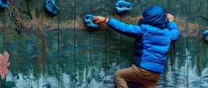 Ein junger Junge klettert an einer blauen Kletterwand mit blauen Handgriffen. Er trägt eine blaue dicke Jacke und braune Hosen.