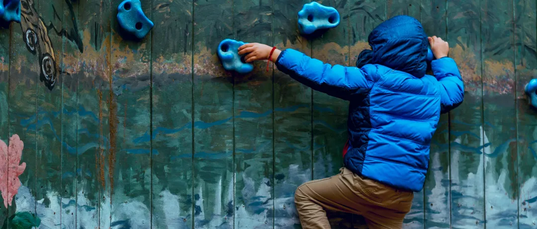 Ein junger Junge klettert an einer blauen Kletterwand mit blauen Handgriffen. Er trägt eine blaue dicke Jacke und braune Hosen.
