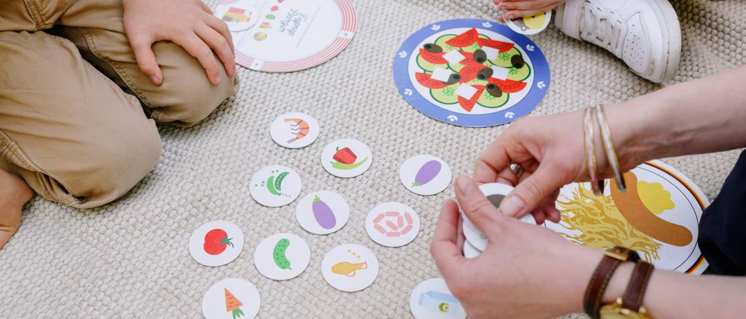 Kinder sitzen auf einem Teppich und spielen ein Essensthematisches Spiel mit runden Lebensmitteln. Sie passen Lebensmittelstücke zu Tellern.