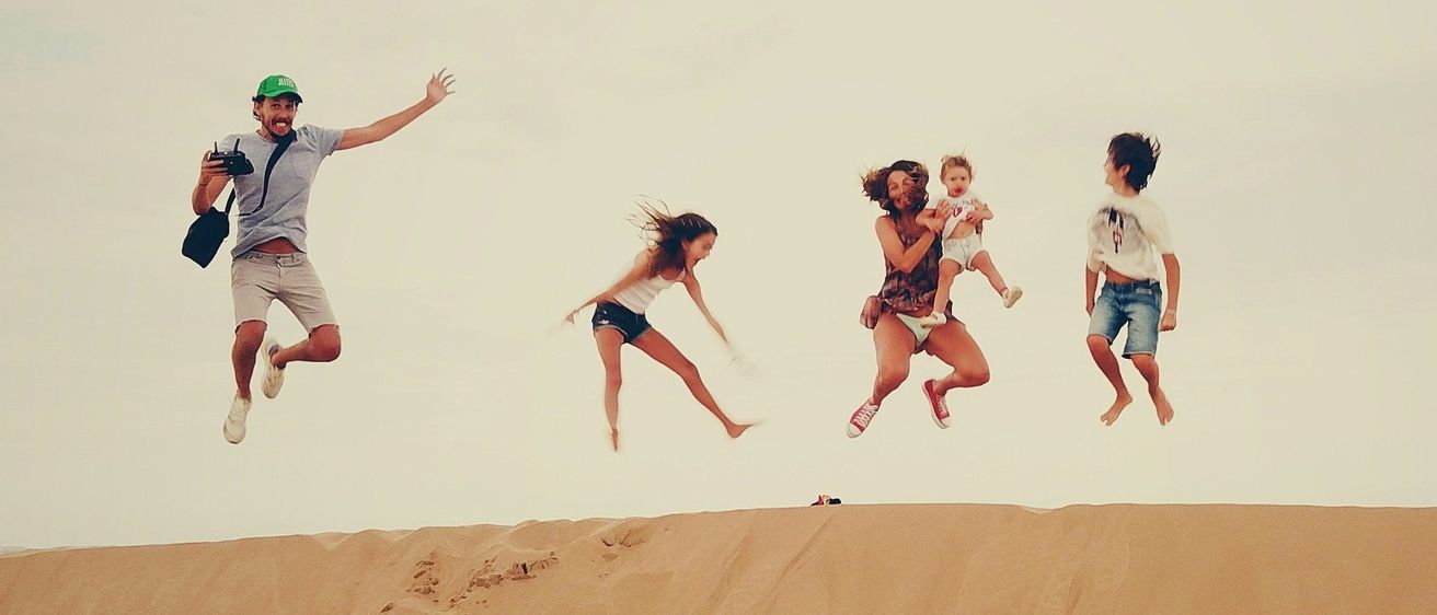 Eine Gruppe von Menschen springt gemeinsam auf einer Düne, unter einem blauen Himmel mit verstreuten Wolken. Einer hält eine Kamera, während andere ein Baby und Kinder tragen.