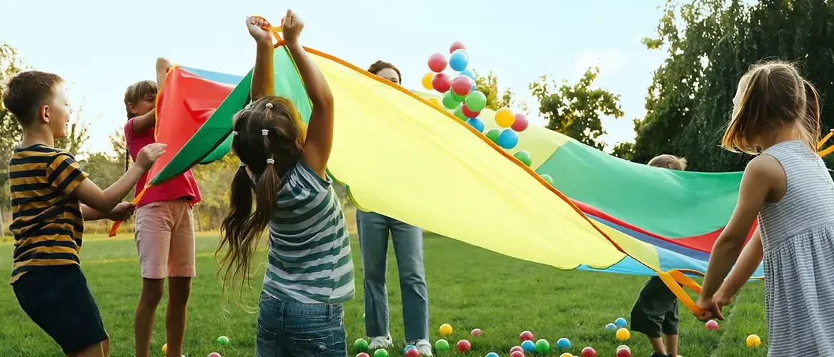 Ein junges Mädchen spielt mit einem großen, farbenfrohen Fallschirm, während eine Frau und ein Kind mit Luftballons auf einer Wiese spielen.