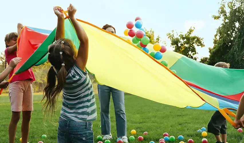 A young girl plays with a large, colorful parachute, while a woman and child play with balloons in a grassy area.