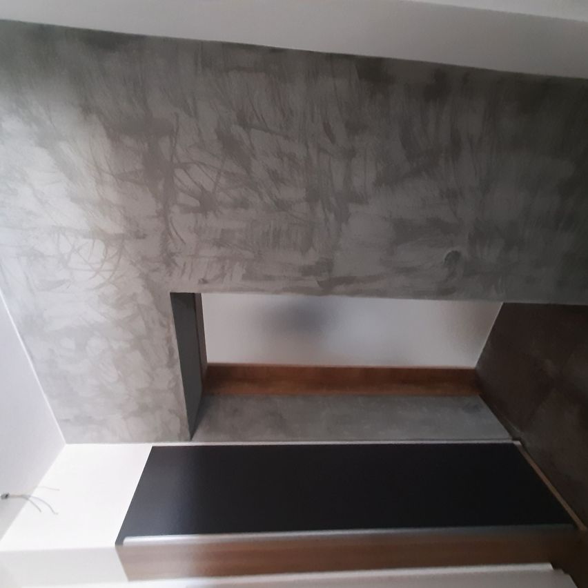 Eine Ecke eines Innenraums mit einer grauen Betonwand und einem Holzboden, mit einem schwarzen Tisch mit Holzfuß.
