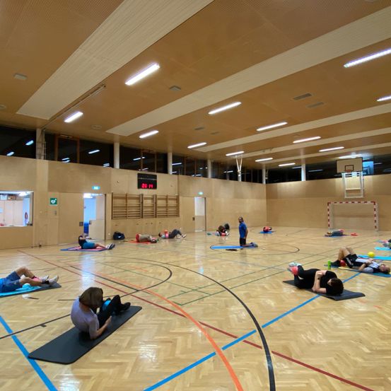 Eine Gruppe von Menschen macht Yoga auf Matten in einer großen Indoor-Sporthalle. Einige liegen, andere stehen. Die Halle hat ein Basketballplatz-Layout.