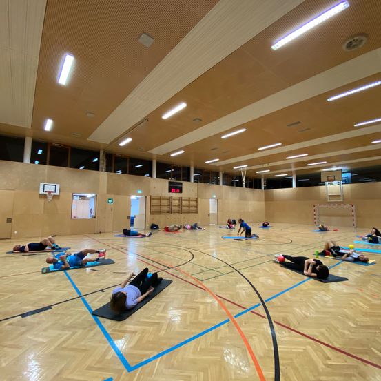 Eine Gruppe von Menschen praktiziert Yoga auf Matten in einer großen Indoor-Sporthalle. Der Boden ist mit blauen und orangefarbenen Linien markiert.