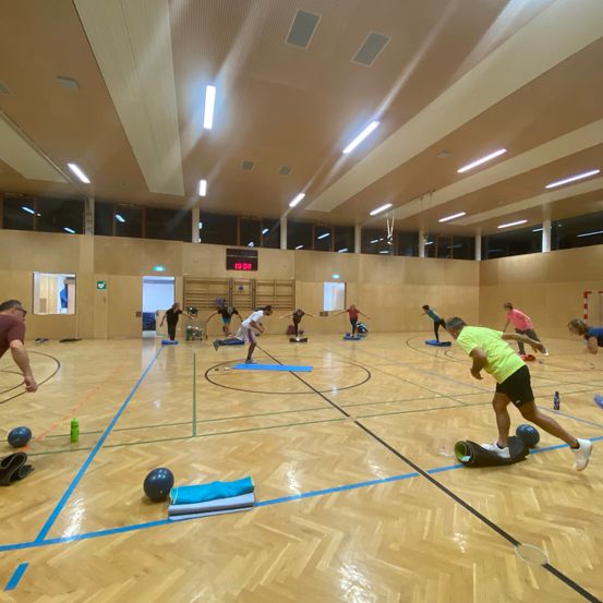 Eine Gruppe von Menschen trainiert in einer Indoor-Sporthalle, einige auf Matten, andere auf Balancebrettern, mit verschiedenen Trainingsgeräten verstreut.