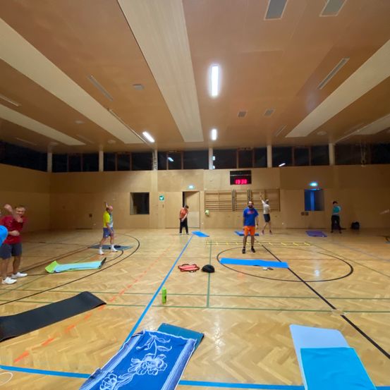 Eine Gruppe von Menschen trainiert in einer Indoor-Sporthalle mit Holzböden und blauen Markierungen. Einige machen Yoga, während andere Trainingsgeräte benutzen.