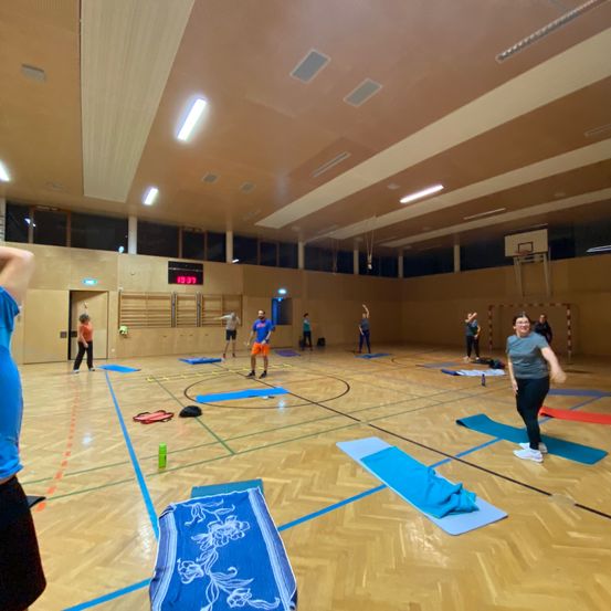 Eine Gruppe von Menschen macht Yoga in einer Turnhalle. Der Raum hat einen Holzboden und einen Basketballkorb. Einige Leute stehen und andere sitzen auf Matten. Es gibt Flaschen und Handtücher auf dem Boden.