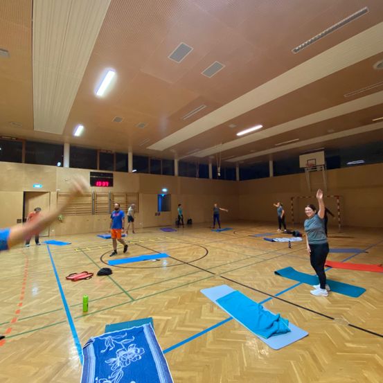 Eine Gruppe von Menschen praktiziert Yoga in einer Turnhalle mit Holzböden, blauen Linien, die den Bereich markieren, und einem Basketballkorb im Hintergrund.