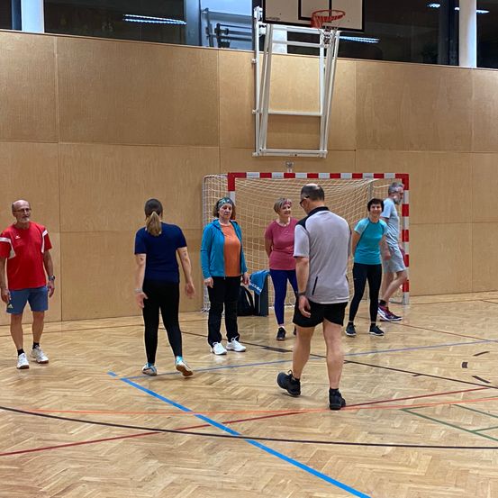 Eine Gruppe von Erwachsenen steht auf einem Basketballplatz, einige tragen Turnschuhe und andere sind barfuß. Sie alle schauen auf etwas vor sich. Der Platz hat einen Basketballkorb und ein Tornetz. Die Wände sind aus Holz.