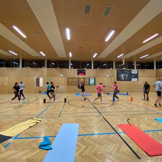 Eine Gruppe von Menschen spielt ein Spiel in einer Indoor-Sporthalle. Sie rennen und springen über Hütchen. Es gibt Yoga-Matten und andere Ausrüstung auf dem Boden. Die Halle hat Holzböden und eine hohe Decke mit Lichtern.