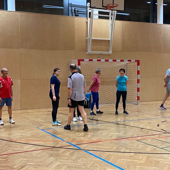 Eine Gruppe von Menschen steht in einer Turnhalle, möglicherweise in Vorbereitung auf ein Spiel. Sie tragen Turnschuhe und einige halten Taschen. Die Halle hat Holzböden und Wände, und es gibt einen Basketballkorb und ein Tor.